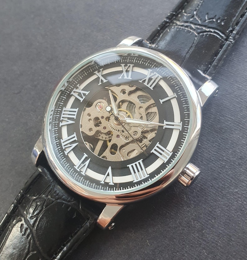 Puede incluir: Un reloj de pulsera plateado con una esfera negra y marcadores de hora en n&uacute;meros romanos. El reloj tiene una parte posterior transparente que revela el funcionamiento interno del movimiento mec&aacute;nico. El reloj est&aacute; en una correa de cuero negra.