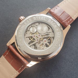 Pode incluir: Um rel&oacute;gio dourado com uma pulseira de couro marrom. A parte de tr&aacute;s do rel&oacute;gio &eacute; transparente, revelando o movimento mec&acirc;nico no interior. O mostrador do rel&oacute;gio &eacute; gravado com "STAINLESS STEEL BACK - MECHANICAL".