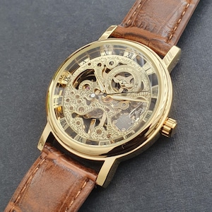 Könnte beinhalten: Goldfarbene Skelettuhr mit braunem Lederarmband. Das Zifferblatt der Uhr zeigt römische Ziffern und komplizierte Innenmechanismen. Das Armband ist in einem satten Braunton gehalten, und die Uhr hat ein klassisches, elegantes Design.