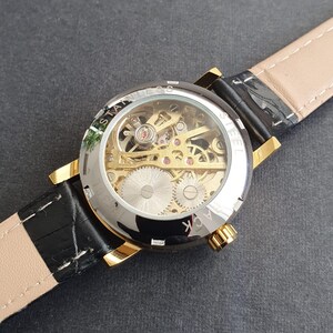 May include: A gold-tone watch with a black leather band. The watch face is clear and shows the inner workings of the watch mechanism. The text "STAINLESS STEEL BACK" is visible on the watch face.