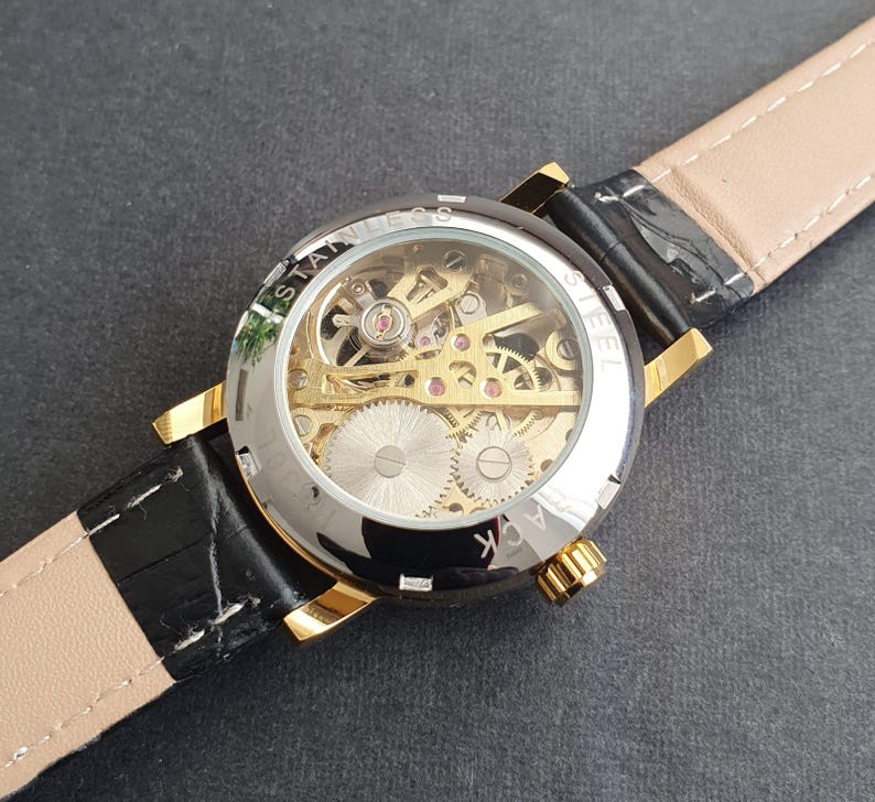 May include: A gold-tone watch with a black leather band. The watch face is clear and shows the inner workings of the watch mechanism. The text "STAINLESS STEEL BACK" is visible on the watch face.
