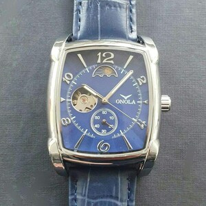 Könnte beinhalten: Eine silberfarbene Armbanduhr mit blauem Zifferblatt, rechteckigem Gehäuse und blauem Lederarmband. Die Uhr verfügt über eine Mondphasenanzeige und ein offenes Herzdesign, das das mechanische Uhrwerk freilegt. Das Zifferblatt der Uhr enthält außerdem ein kleines Sekunden-Zifferblatt und den Markennamen "ONOLA", der auf dem Zifferblatt aufgedruckt ist.