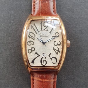 Puede incluir: Un reloj de pulsera dorado con una correa de cuero marrón. La esfera del reloj es de color crema con grandes números negros y la marca "Chenxi". El reloj tiene una pantalla de fecha a las 6 en punto.