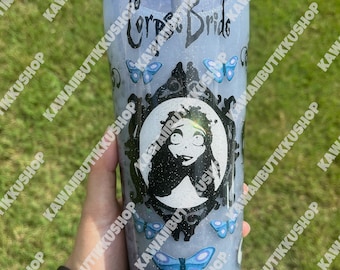 May include: A clear tumbler with a black straw, featuring a design from the movie "Corpse Bride." The tumbler has a glittery gray background with blue butterflies and a skull. The top rim is decorated with black and iridescent rhinestones.
