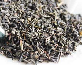 May include: Close-up of loose leaf green tea spilling from a white bowl. The tea leaves are a mix of dark and light green, with a slightly curled texture. The text "Le Virg" is visible in the image.