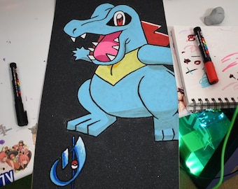 May include: A black skateboard deck with a blue and yellow cartoon drawing of a Pokemon character. The character is a blue lizard with a yellow collar and red accents. The text "PanchoVillao2022" is written in black at the bottom of the deck.