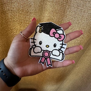 May include: White embroidered patch of Hello Kitty wearing a graduation cap and holding a diploma. The cap is black with a gold tassel. The diploma is white with a pink ribbon.