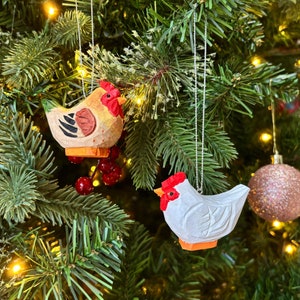 May include: Two wooden chicken ornaments hanging on a Christmas tree. One chicken is white and the other is yellow with brown and red accents.
