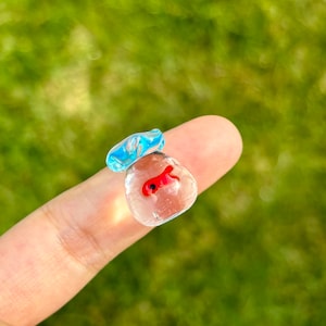 May include: A small glass figurine of a goldfish in a clear glass bag with a light blue top.