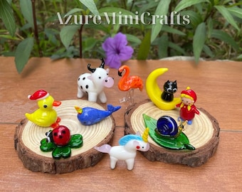 May include: A collection of miniature glass animal figurines including a yellow duck, a spotted cow, a pink flamingo, a blue whale, a black cat on a yellow moon, a red ladybug, a unicorn, and a snail on a green leaf, displayed on wood slices.