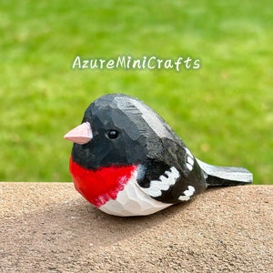 May include: Hand-carved wooden bird figurine painted in black, white, and red. The bird has a black head, white body, and red breast. The bird is sitting on a brown surface.