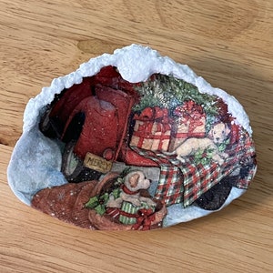 May include: A white ceramic seashell with a snowy rim, decorated with a Christmas scene featuring a red truck carrying a Christmas tree and presents, two dogs, and the word "Merry".