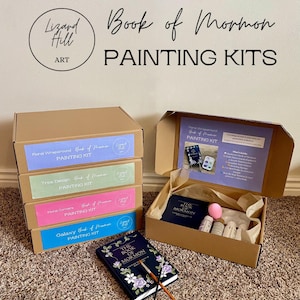 Puede incluir: Kits de pintura "Book of Mormon" en cajas de cartón, una abierta mostrando contenido como un libro y pintura. El texto "Lizard Hill Art" y "PAINTING KITS" es visible. Sobre una alfombra.