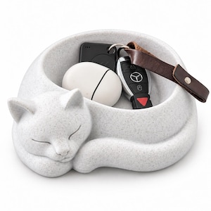 Puede incluir: Un cuenco de color gris claro moteado con forma de gato y acabado mate liso. El gato aparece en posición acurrucada y durmiendo, con la cabeza apoyada sobre las patas. Dentro del cuenco hay un estuche ovalado blanco para auriculares y una llave.
