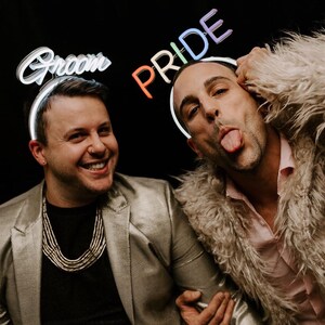 May include: Two people wearing neon signs that say "Groom" and "Pride" on their heads. The person on the left is wearing a silver jacket and a necklace. The person on the right is wearing a furry jacket and sticking their tongue out.