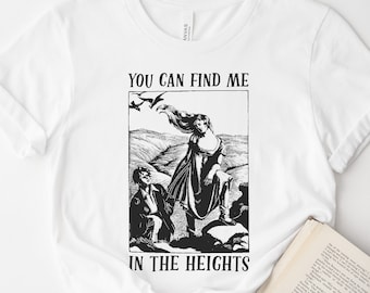 May include: White t-shirt featuring a black and white illustration with the text "YOU CAN FIND ME IN THE HEIGHTS." The graphic depicts a woman standing on a hill with two figures below. A book and a pink rose are in the foreground.