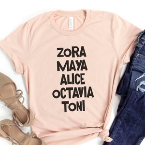 May include: A light pink t-shirt with black text that reads "Zora Maya Alice Octavia Toni". The shirt is paired with blue jeans and brown shoes.