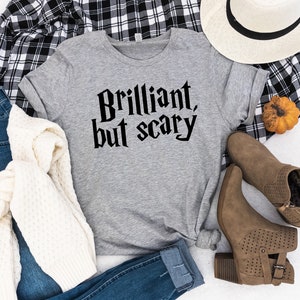 May include: A gray t-shirt with the text "Brilliant, but scary" printed in black. The shirt is laid out on a white surface with a plaid shirt, a white hat, a pair of brown boots, a white sweater, and a pair of blue jeans.