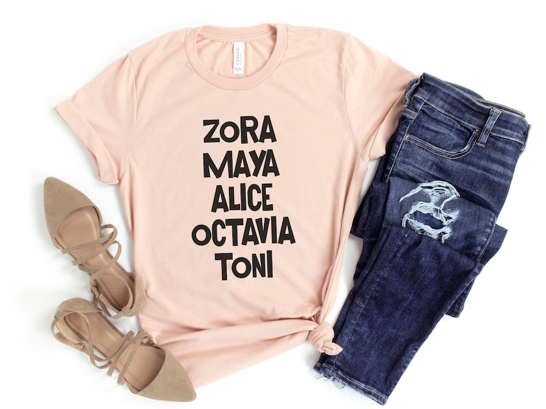 May include: A light pink t-shirt with black text that reads "Zora Maya Alice Octavia Toni". The shirt is paired with blue jeans and brown shoes.