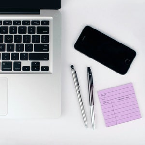May include: A silver laptop computer with a black keyboard and trackpad, a black smartphone, two silver pens, and a purple lined notepad on a white surface.