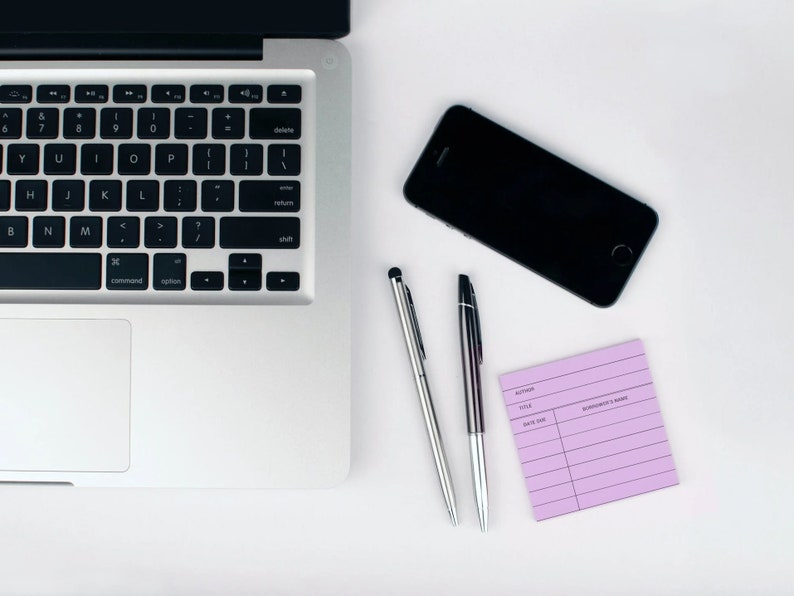 May include: A silver laptop computer with a black keyboard and trackpad, a black smartphone, two silver pens, and a purple lined notepad on a white surface.