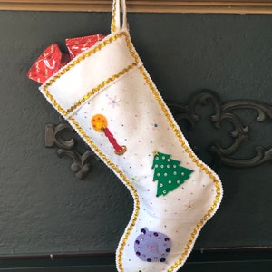 Könnte beinhalten: Weißer Weihnachtssocken mit goldener Einfassung und festlichen Verzierungen, darunter ein grüner Baum, ein violetter Weihnachtsbaumschmuck und eine rot-gelbe Kerze. Der Socken ist mit verpackten Geschenken gefüllt.