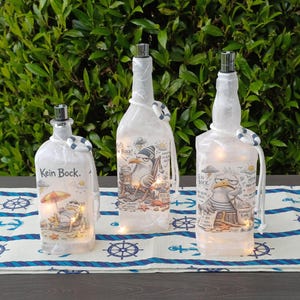Könnte beinhalten: Drei dekorative Glasflaschen mit silbernen Stöpseln und Lichterketten. Jede Flasche zeigt eine nautische Illustration einer Möwe und den Text "Kein Bock". Die Flaschen sind mit kleinen Rettungsringen verziert.