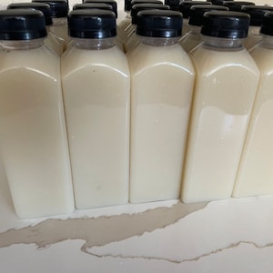 May include: A row of 10 clear plastic bottles with black lids filled with a white liquid. The bottles are arranged in a line on a white surface.