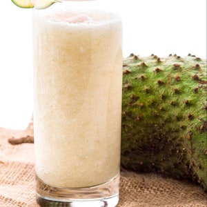 May include: A tall glass of creamy, light-colored smoothie with a lime wedge garnish. The smoothie is sitting on a brown burlap surface next to a green, spiky soursop fruit.