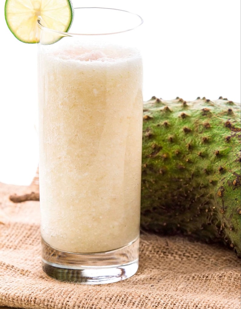 May include: A tall glass of creamy, light-colored smoothie with a lime wedge garnish. The smoothie is sitting on a brown burlap surface next to a green, spiky soursop fruit.