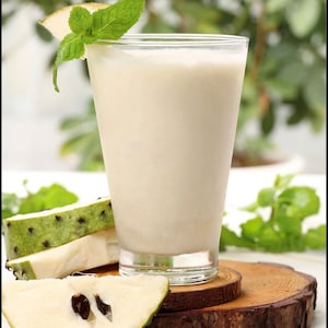 May include: A glass of creamy white smoothie with a sprig of mint on the rim, sitting on a wooden coaster with slices of green and white fruit.