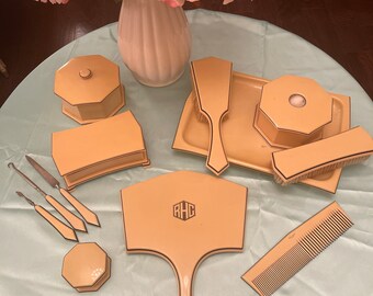 Può includere: Un set da toeletta vintage con uno specchio da mano, un pettine, spazzole e piccole scatole. Il set è realizzato in un materiale giallo pallido con finiture nere e le iniziali "RHC" sullo specchio.