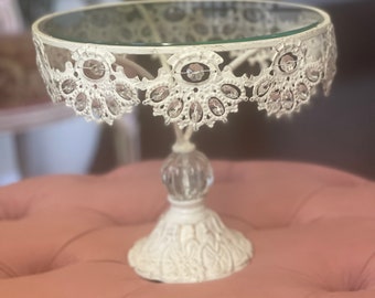 May include: White metal cake stand with a glass top and crystal accents. The stand has a scalloped edge and a decorative base.