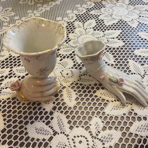 May include: Two white porcelain vases with floral designs. One vase is shaped like a hand with pink flowers and the other is a traditional vase with a hand holding it.
