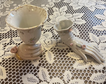 Könnte beinhalten: Zwei weiße Porzellanvasen mit Blumendesigns. Eine Vase ist handförmig mit rosa Blumen und die andere ist eine traditionelle Vase, die von einer Hand gehalten wird.