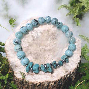 May include: A beaded bracelet featuring light blue and black patterned round beads, interspersed with turquoise-colored irregular beads and silver-toned accents. The bracelet is displayed on a wooden surface.