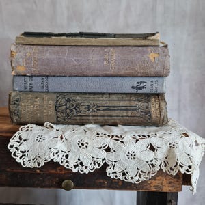 May include: Four vintage books with aged covers in grey, purple, and brown tones, stacked on a wooden surface. A white lace doily is beneath the books. The titles and authors are visible on the spines of the books.