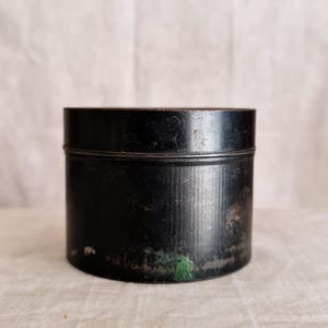 May include: A small, round, black metal container with a lid. The container has a cylindrical shape and shows signs of age, with some discolouration and wear visible. The background is a light, neutral colour.