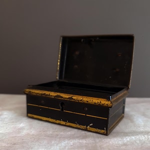 May include: An open, antique black metal box with gold trim and accents. The box has a keyhole and is sitting on a white surface. The interior of the box is black.