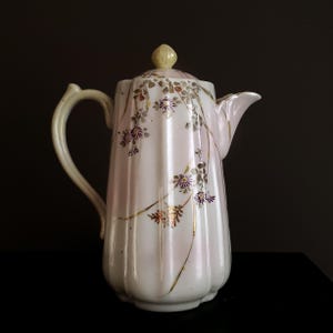 May include: A white porcelain coffee pot with a lid, handle, and spout. The pot is decorated with pink and gold floral designs. The handle and lid finial are a pale yellow. The pot is on a black surface.