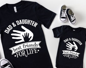 May include: Black t-shirts and a baby onesie with the text "DAD & DAUGHTER best friends FOR LIFE" in white. The design includes a graphic of two hands, one large and one small. A pair of black sneakers and baby shoes are also in the image.