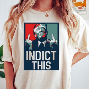 May include: A white t-shirt with a graphic of a man giving the middle finger with the text "Indict This" in red, white, and blue.