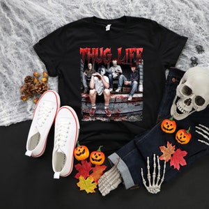May include: Black t-shirt with the text "THUG LIFE" in red, featuring a graphic of four people. White trainers, blue jeans, a skull, and pumpkins are arranged around the shirt, creating a Halloween theme.