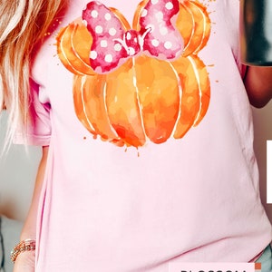 Op de afbeelding: Lichtroze T-shirt met een aquarelontwerp van een oranje pompoen en een roze strik met stippen. Het woord "BLOSSOM" is gedrukt in een witte rechthoek aan de onderkant. Het shirt heeft korte mouwen.