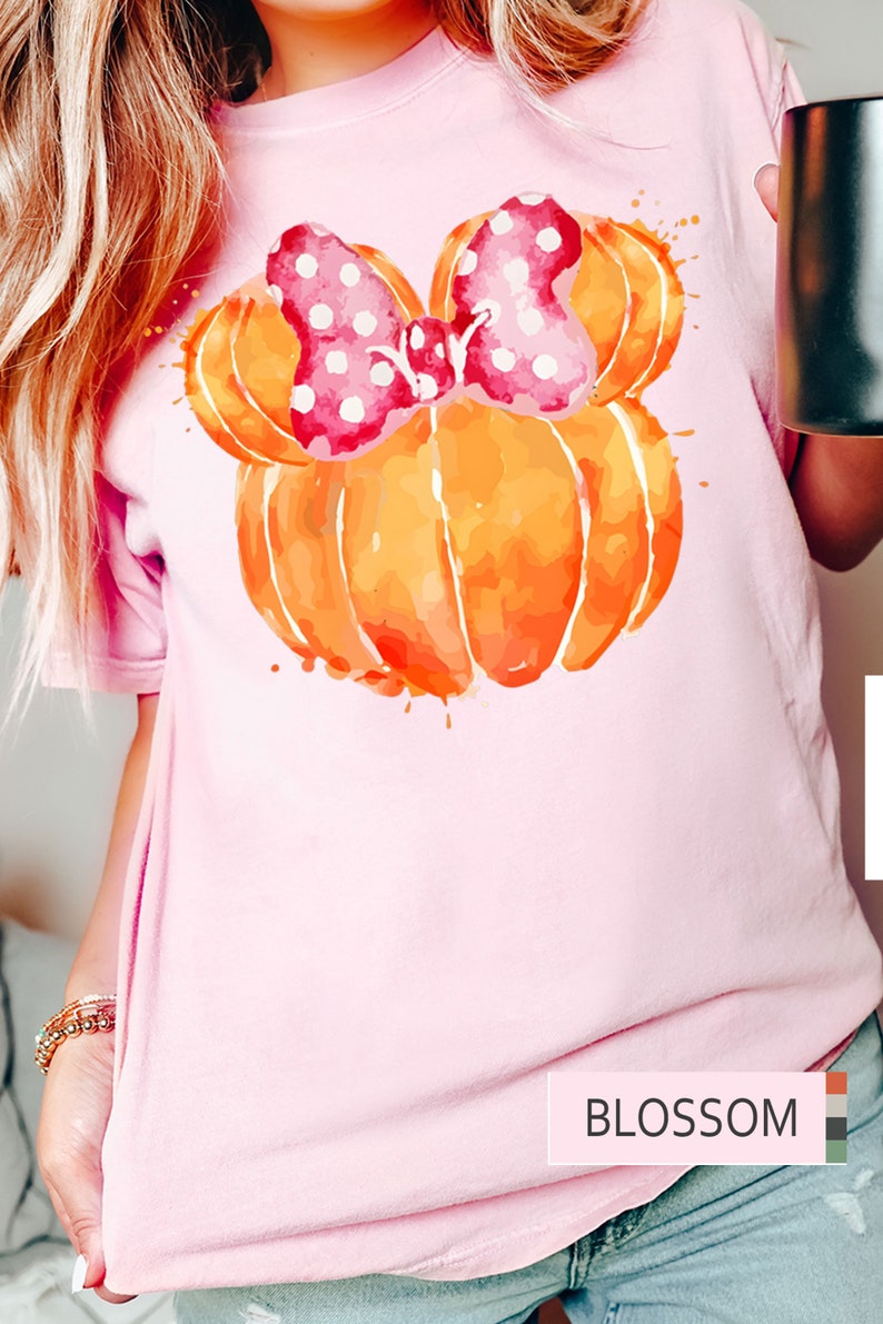 Op de afbeelding: Lichtroze T-shirt met een aquarelontwerp van een oranje pompoen en een roze strik met stippen. Het woord "BLOSSOM" is gedrukt in een witte rechthoek aan de onderkant. Het shirt heeft korte mouwen.