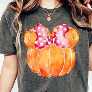 Op de afbeelding: Donkergrijs T-shirt met een aquarelontwerp van twee oranje pompoenen en een roze strik met stippen. Het woord "PEPPER" staat onderaan gedrukt. Het shirt heeft een casual stijl.