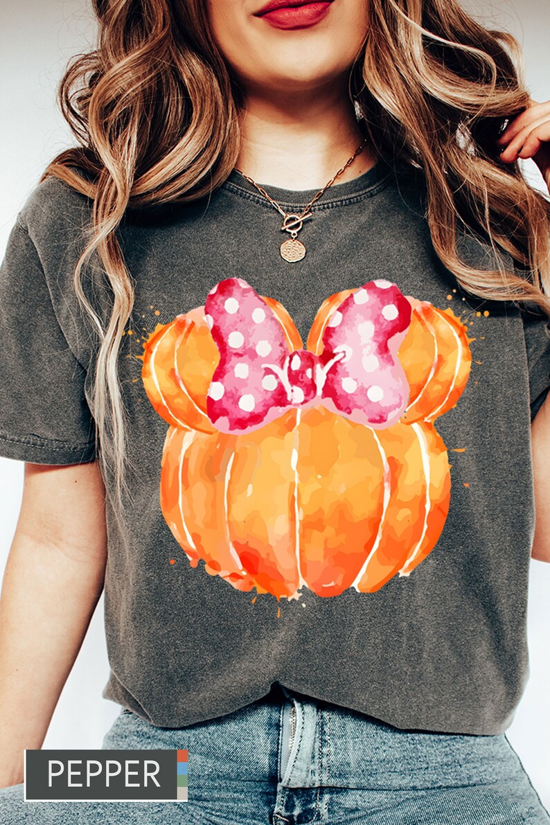 Op de afbeelding: Donkergrijs T-shirt met een aquarelontwerp van twee oranje pompoenen en een roze strik met stippen. Het woord "PEPPER" staat onderaan gedrukt. Het shirt heeft een casual stijl.