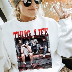May include: White crew neck sweatshirt with a graphic of horror movie characters and the text "THUG LIFE" in red. The design includes Jason Voorhees, Michael Myers, Pennywise, and Freddy Krueger. The sweatshirt is a casual style.