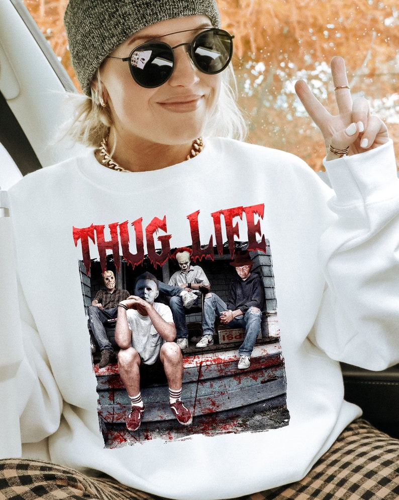 May include: White crew neck sweatshirt with a graphic of horror movie characters and the text "THUG LIFE" in red. The design includes Jason Voorhees, Michael Myers, Pennywise, and Freddy Krueger. The sweatshirt is a casual style.