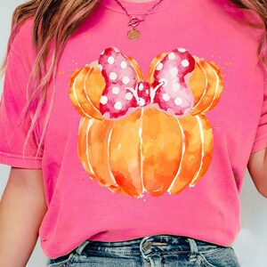 Op de afbeelding: Felroze T-shirt met een aquarelontwerp van een pompoen en een roze stippenstrik. De pompoen is oranje met een aquareleffect. Het shirt heeft een ronde hals.
